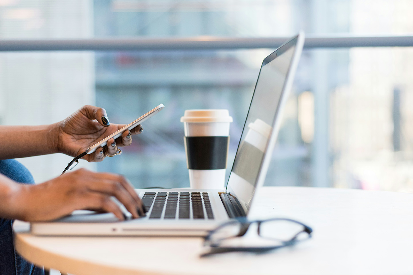 woman typing on a laptop with one hand, holding a phone in the other at a desk with a coffee in the background