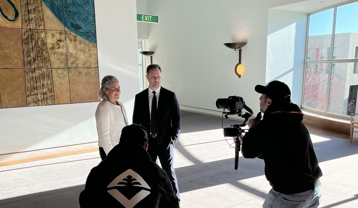 Rom and Damian standing infront of an abstract artwork in a large room with two cameramen pointing equipment at them