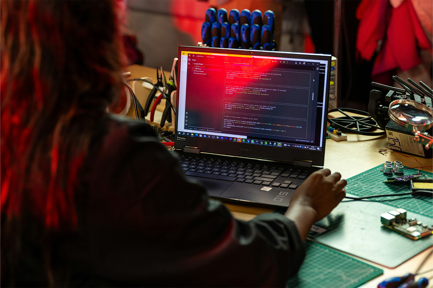 Woman typing on a laptop with code on it, mechanical equipment lying on the desk around the laptop