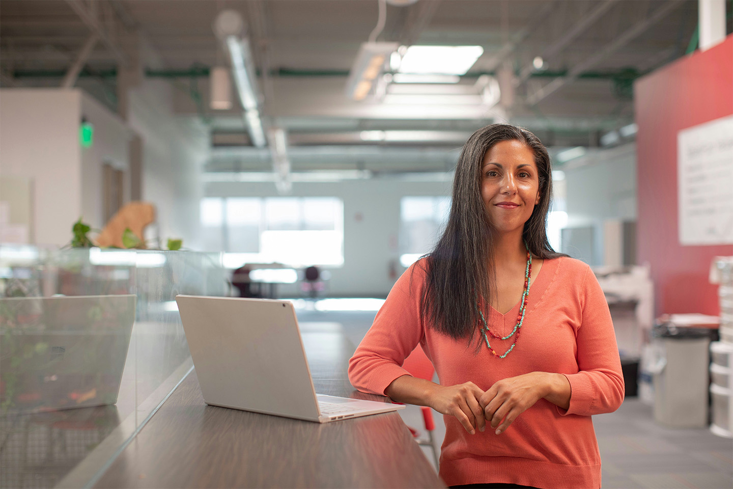 woman standing next to a laptop on her desk, wearing an orange shirt