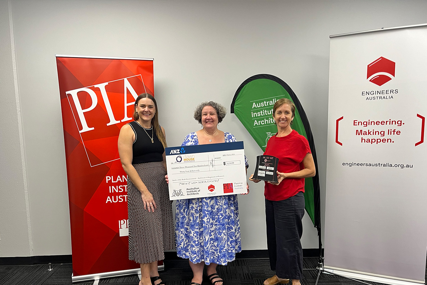Three people stand in front of company banners, the woman in the middle is holding an oversized cheque