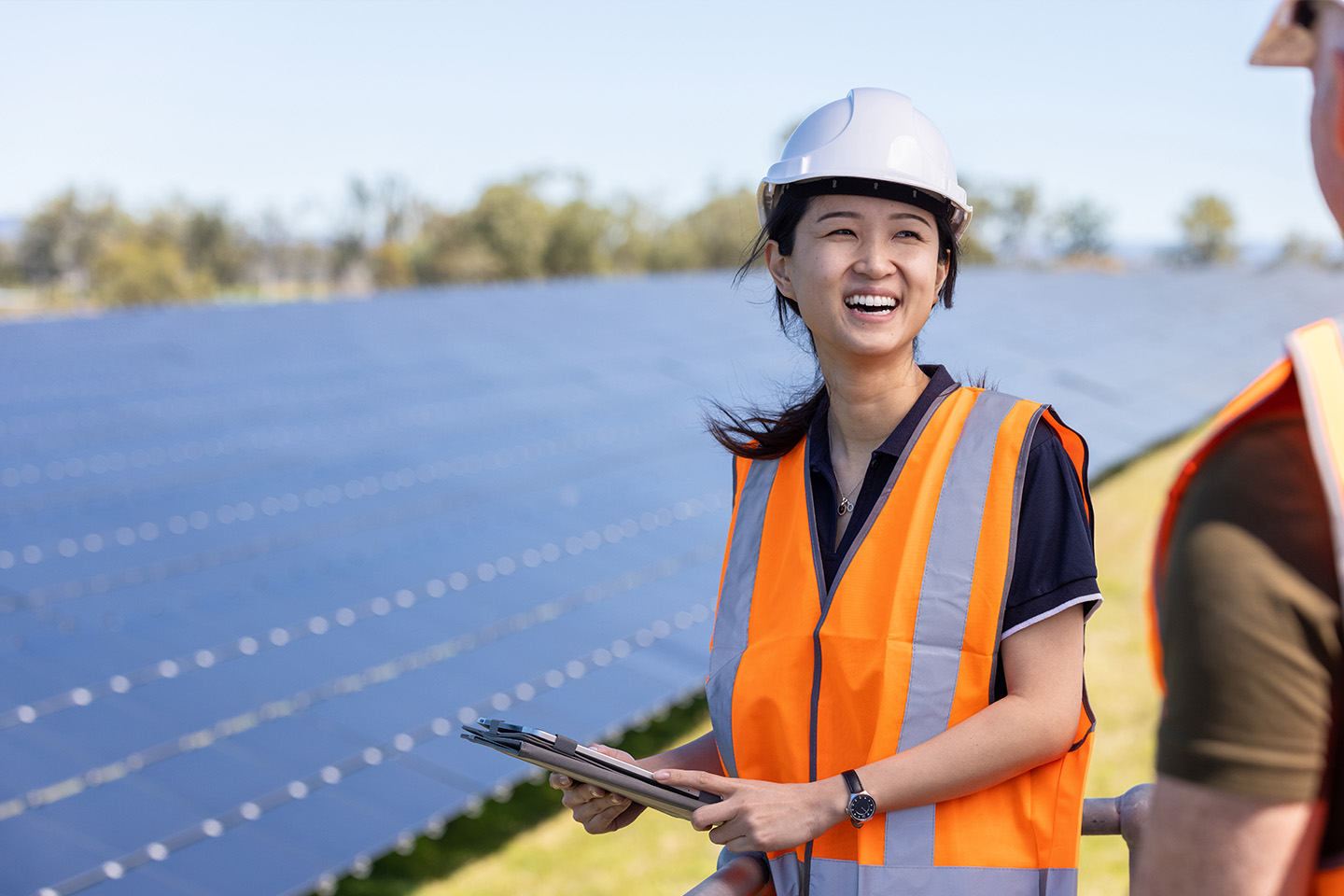 Woman with orange safety vest and white hard hat on standing in front of a large line of solar panels