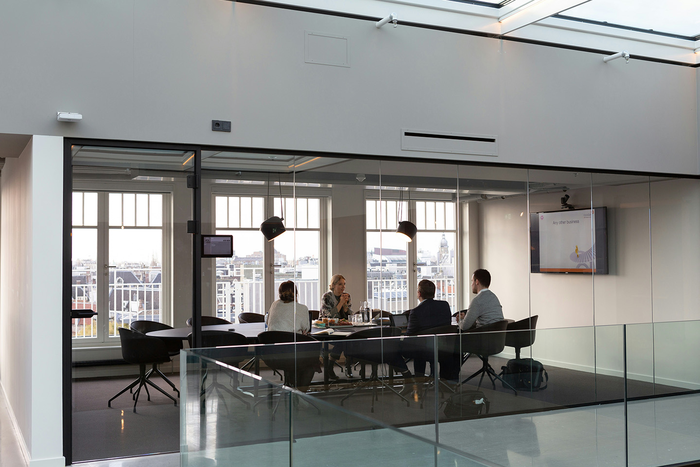 Large meeting room with floor to ceiling windows, four people sat at the desk inside the meeting room chatting