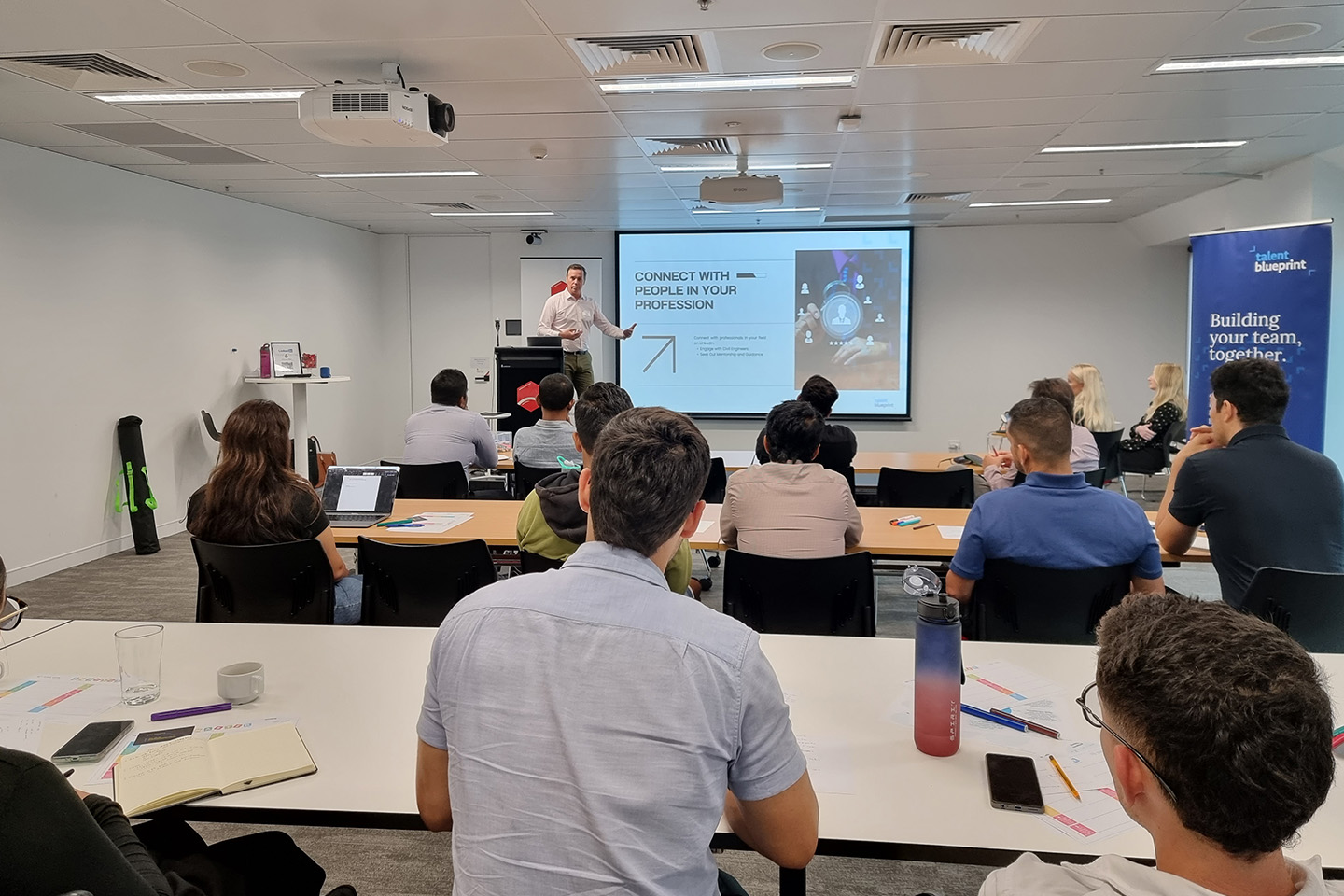 People at desks in a large room looking toward someone presenting at the front of the room next to a projector