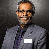 Image of a man in a grey suit and glasses smiling at the camera, dark backdrop
