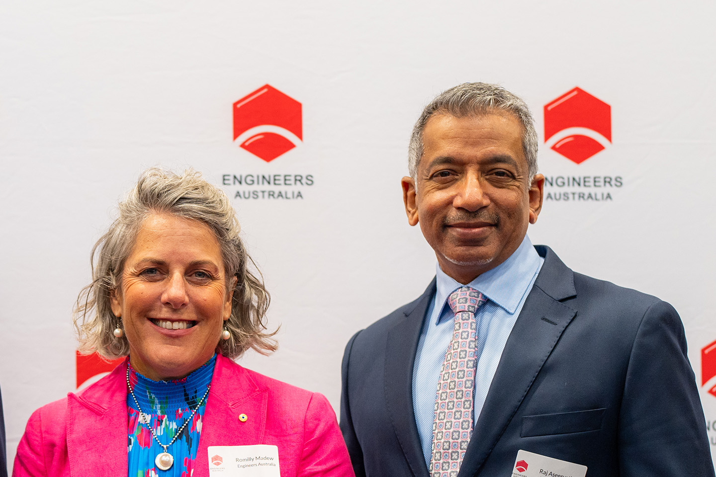 Romilly smiling in a pink blazer and blue top next to Raj also smiling in a grey suit and blue shirt