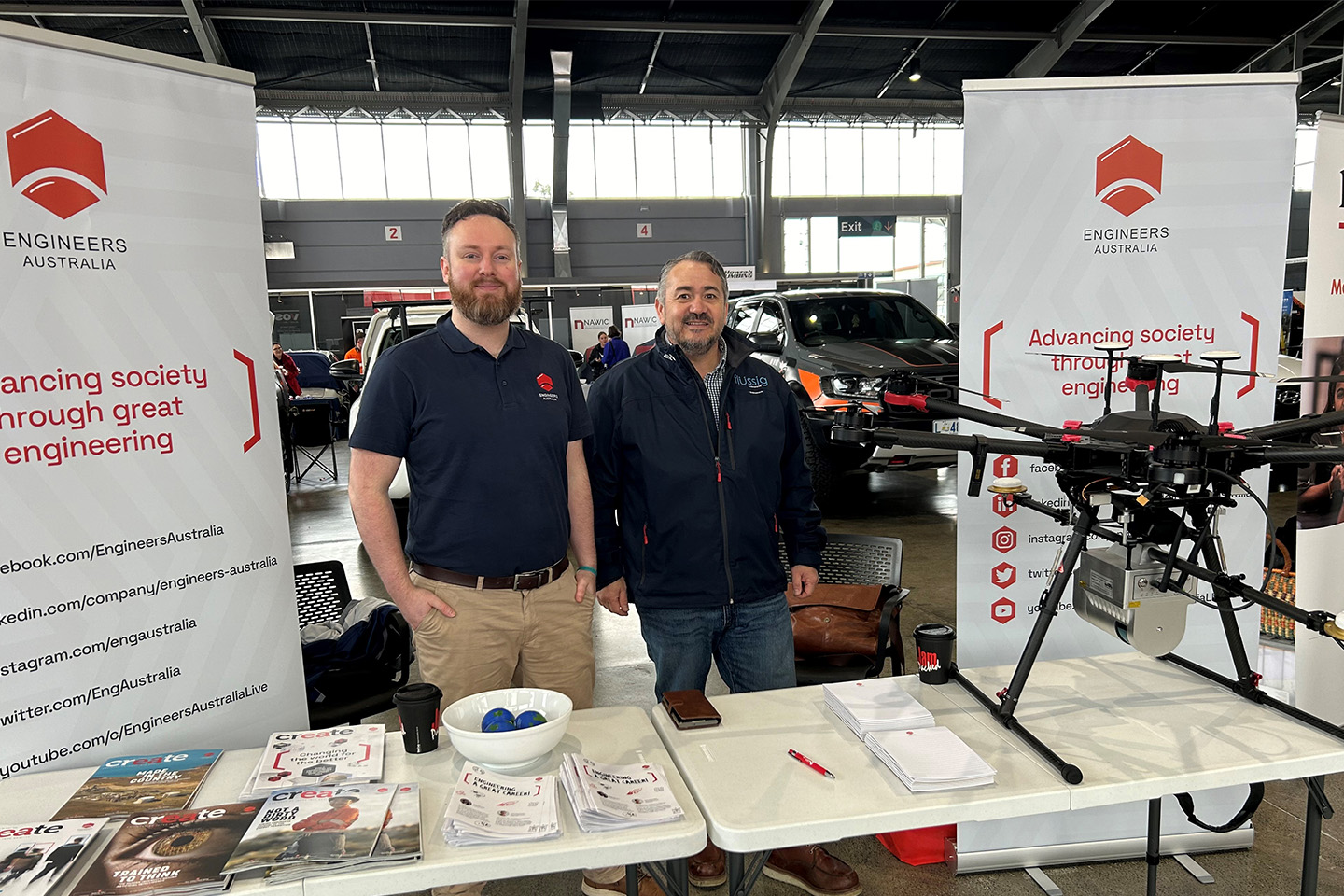Two men standing behind a booth with a large electronic drone in front of them on a table