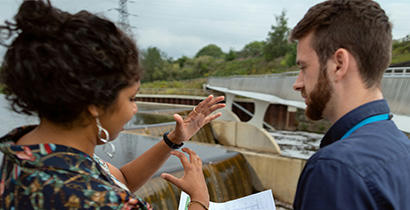 Woman and a man talking over printed plans in front of a stream of water